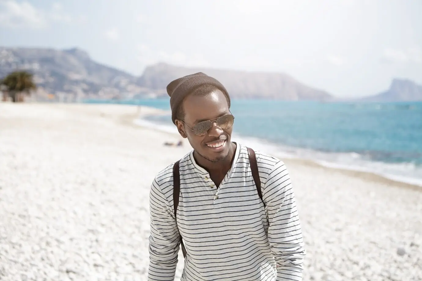 Close-up portrait of a laughing young, handsome, dark-skinned backpacker wearing mirrored-lens sunglasses and a hat, feeling happy while walking down the beach on a summer weekend.
