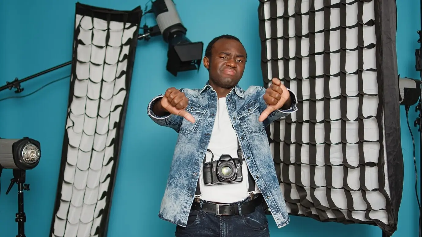 A young man giving a thumbs-down gesture of dislike in a studio with professional photography equipment, expressing disagreement and disapproval, with a tripod and backstage lighting.