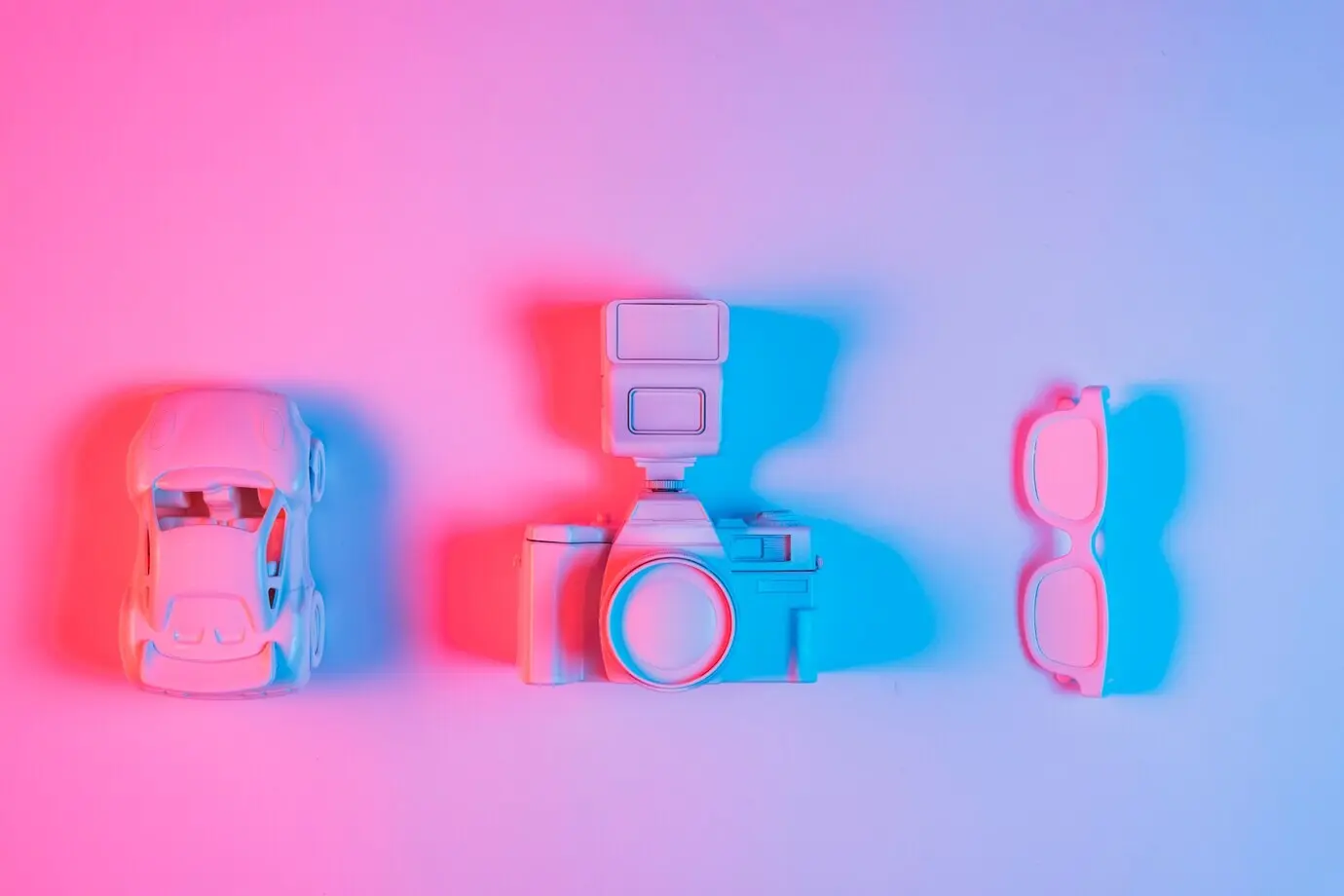 A toy car, a retro camera, and spectacles arranged in a row on a pink backdrop with a blue light effect.