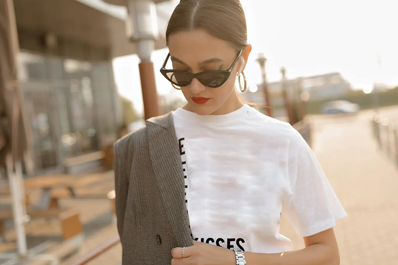 Portrait of a stylish woman in sunglasses walking down the street; she is wearing a white T-shirt, has red lips, and is looking down.