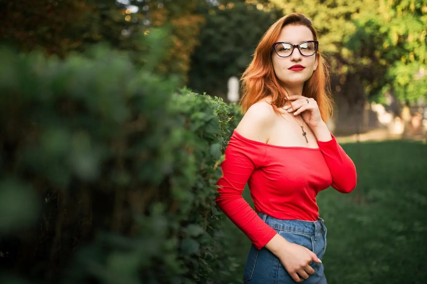 An attractive red-haired woman wearing eyeglasses, a red blouse, and a jeans skirt posing in a green park.